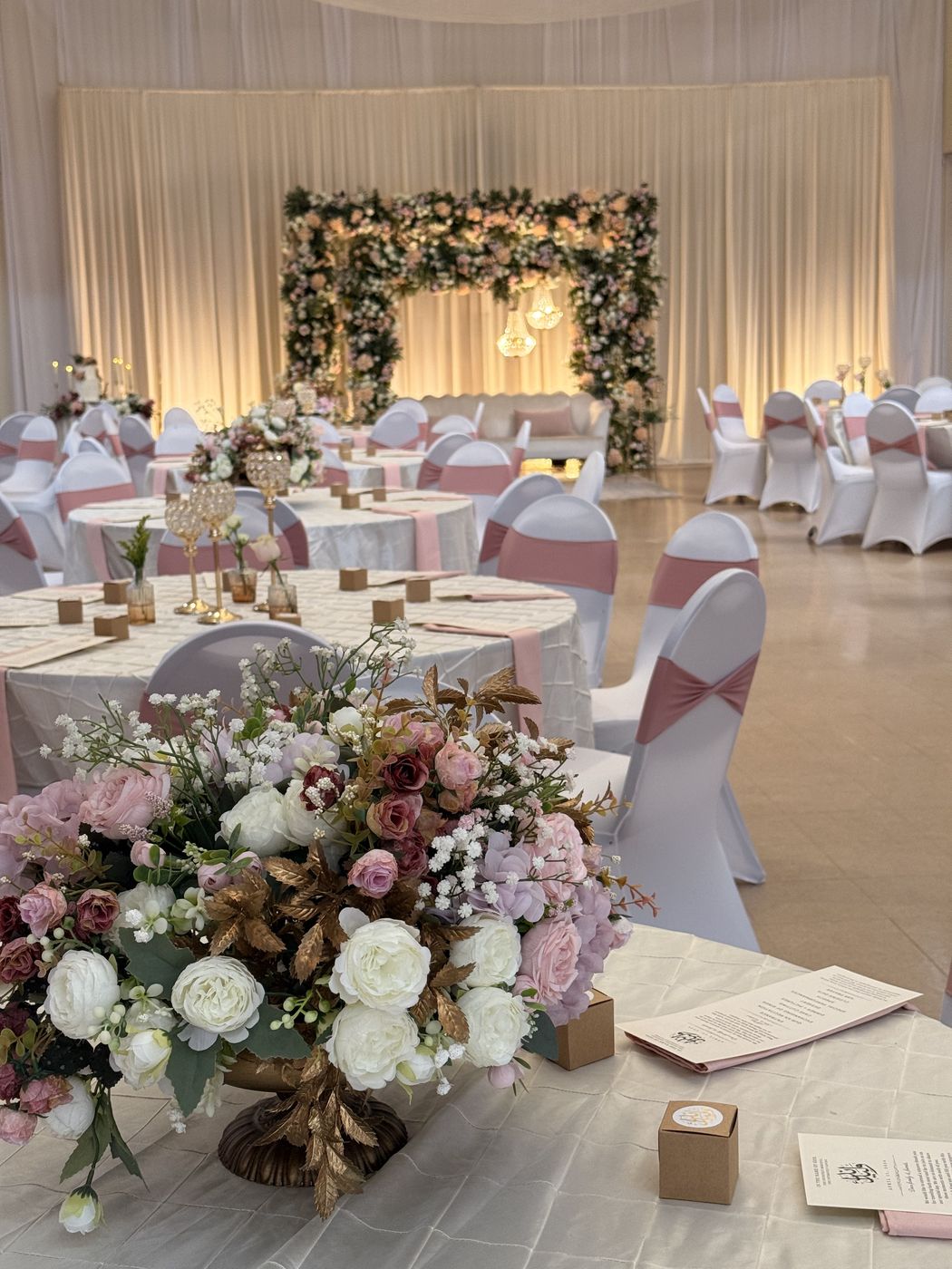 Round tables with floral centerpieces and a flower arch backdrop set for a wedding reception